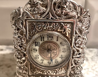 Könnte beinhalten: Eine silberne Uhr mit einem verzierten, detaillierten Design. Das Zifferblatt der Uhr hat römische Ziffern und ein zartes Blumenmuster.