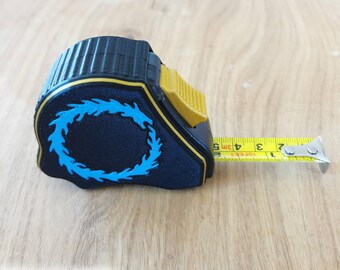 May include: A black tape measure with a yellow locking switch and a blue circular flame-like graphic on the side. The extended yellow metal tape shows markings in both inches and centimeters, with the text 10 FEET and 3M clearly visible on the blade.
