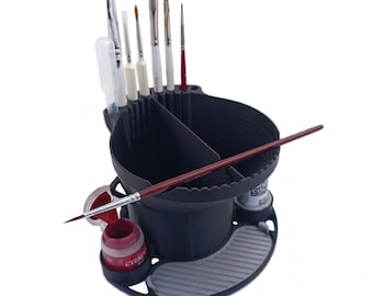 May include: A black plastic paint brush and water cup organizer featuring multiple vertical slots for brushes, a central divided basin, a red paint pot, a grey textured cleaning pad, and the text PRODICER printed at the bottom left corner.