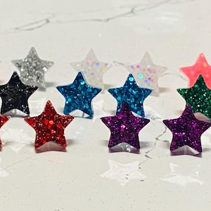 May include: A set of 10 star-shaped earrings with glitter. The earrings are in various colors, including red, blue, green, purple, pink, and silver.