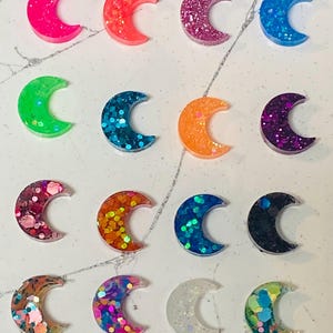 May include: Sixteen crescent moon-shaped resin charms arranged in a four-by-four grid. Each piece features a unique color and glitter finish, including neon pink, orange, purple, blue, green, black, and multi-colored confetti-style glitter patterns.