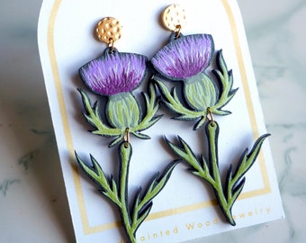 May include: A pair of hand-painted wooden earrings featuring purple and green thistle flowers. The earrings are attached to gold-toned posts with textured tops. The card reads "Golden Fern Adornments Hand-painted Wood Jewelry".