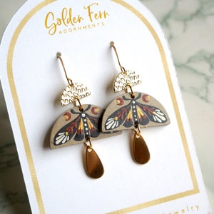 Puede incluir: Pendientes de madera pintados a mano con diseño de polilla. Los pendientes tienen ganchos dorados, detalles de semicírculo y colgantes en forma de lágrima. El diseño de la polilla presenta detalles marrones, negros, blancos y dorados. La tarjeta dice "Golden Fern Adornments."