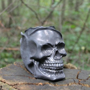 Op de afbeelding: Een zwarte schedel plantenpot gemaakt van keramiek. De schedel heeft een gedetailleerd ontwerp met een gesloten mond en ogen. De pot staat op een boomstronk.