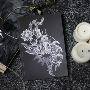 Può includere: Un taccuino nero con un'illustrazione bianca di un occhio circondato da fiori e foglie. Il taccuino è su una superficie grigia con tre candele bianche.