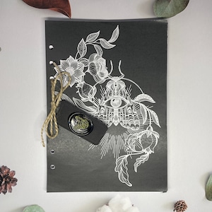 May include: A black journal with a white illustration of a moth with an eye on its wings, surrounded by flowers. The journal has a brown string attached to it.