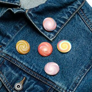 Puede incluir: Cinco botones redondos con diseños de remolinos coloridos están colocados sobre una chaqueta vaquera azul. Los botones presentan un patrón en espiral en tonos de amarillo, rosa y azul. La chaqueta tiene cierre de botón y cuello.