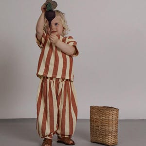 Peut inclure: Un ensemble deux pièces composé d'un haut à manches courtes et d'un pantalon ample, tous deux dotés d'un motif à rayures verticales orange rouille et crème. L'ensemble est assorti de sandales en cuir marron et d'un panier en osier tressé.