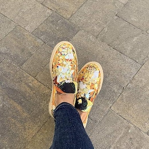 May include: A pair of slip-on shoes with a vibrant, ornate design in shades of gold, yellow, and white. The shoes have a woven, tan-colored sole and black accents. The shoes are worn with blue denim jeans.