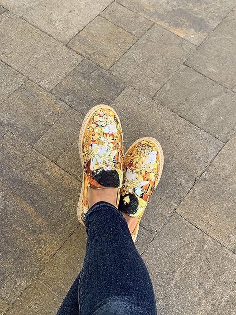 May include: A pair of slip-on shoes with a vibrant, ornate design in shades of gold, yellow, and white. The shoes have a woven, tan-colored sole and black accents. The shoes are worn with blue denim jeans.