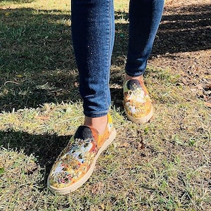 May include: A person wearing blue jeans and colorful slip-on shoes with floral and abstract designs. The shoes have a woven jute sole and a black heel. The shoes are on a grassy surface.