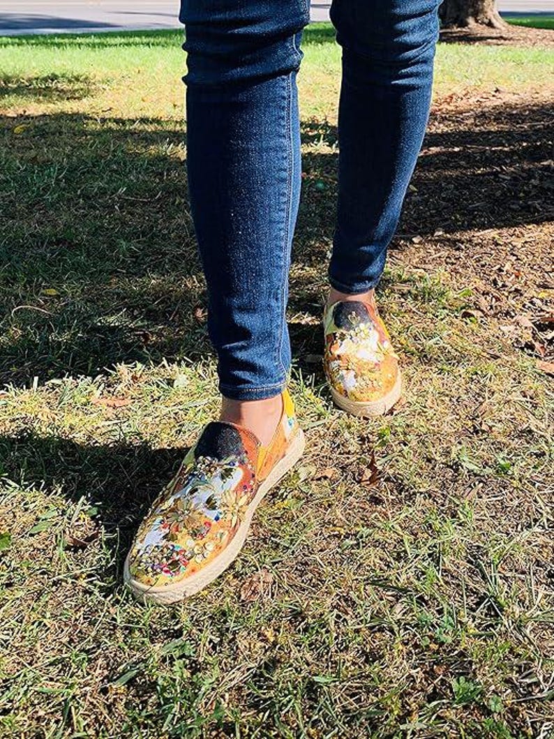 May include: A person wearing blue jeans and colorful slip-on shoes with floral and abstract designs. The shoes have a woven jute sole and a black heel. The shoes are on a grassy surface.