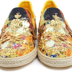 May include: A pair of slip-on shoes with a colorful floral pattern and gold sequin embellishments. The shoes have a yellow heel and a beige rope sole. The design features a mix of orange, yellow, and blue hues.