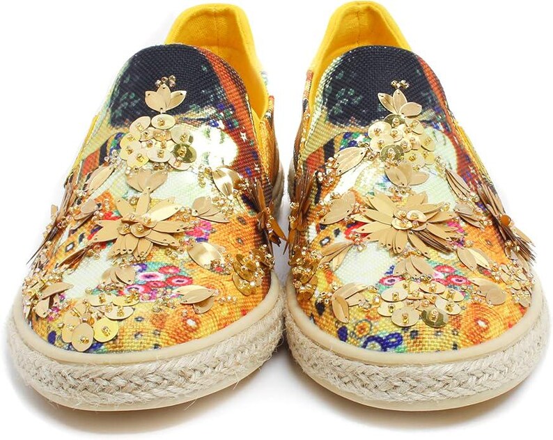 May include: A pair of slip-on shoes with a colorful floral pattern and gold sequin embellishments. The shoes have a yellow heel and a beige rope sole. The design features a mix of orange, yellow, and blue hues.