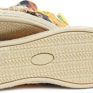 May include: Close-up of the soles of two espadrille shoes. The soles are beige with a textured pattern. The upper part of the shoes features a colorful floral design with yellow, orange, and black accents.