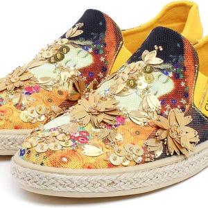 May include: A pair of slip-on canvas shoes with a colorful floral and abstract design. The shoes are embellished with gold sequins and flower appliqu&eacute;s. The soles are made of woven rope, and the interior is yellow. The text "CVITAV KLIMT" is printed on the side.