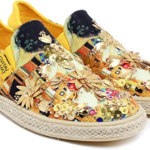 May include: Slip-on canvas shoes with a colorful, abstract design, embellished with gold sequins and floral accents. The shoes feature a yellow heel and a woven sole. The text "GUSTAV KLIMT" is printed on the side.