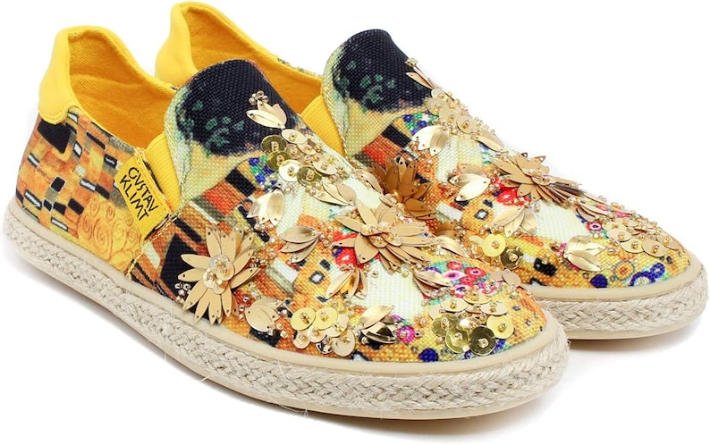 May include: Slip-on canvas shoes with a colorful, abstract design, embellished with gold sequins and floral accents. The shoes feature a yellow heel and a woven sole. The text "GUSTAV KLIMT" is printed on the side.