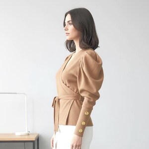 Könnte beinhalten: Eine Frau trägt ein braunes Wickeloberteil mit Puffärmeln und goldenen Knöpfen, kombiniert mit einem weißen Bleistiftrock und beigen Absätzen. Das Outfit ist im Business-Casual-Stil gehalten.