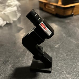 May include: A black laser pointer with a red "DANGER" warning label attached to the side. The laser pointer is mounted on a black stand.