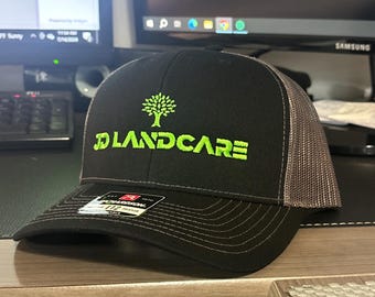 May include: Black and gray trucker hat with a green tree and the text "D LANDCARE" embroidered on the front.