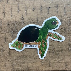 May include: A sticker featuring a sea turtle illustration. The turtle has a dark brown shell, green head and limbs, and brown and tan markings. The text "TRENT CARDINAL ART" is printed on the sticker.