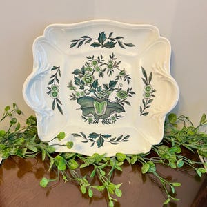 May include: A square, cream-coloured ceramic serving dish with a scalloped edge. The dish features a central floral design in shades of green and dark green, surrounded by smaller floral and leaf motifs. The dish is decorated with a botanical theme.