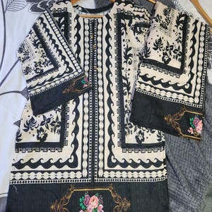 Op de afbeelding: Een tweedelige outfit in crème en zwart, bestaande uit een top en een bijpassende broek. De top heeft een knoopsluiting aan de voorkant en beide stukken zijn versierd met een zwart-crèmekleurig bloemmotief. Bloemenaccenten zijn aanwezig op de randen.