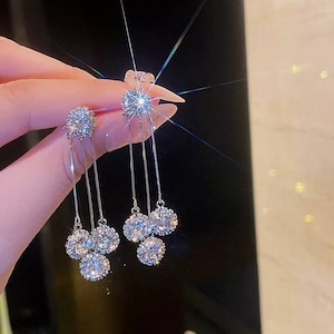 May include: Silver dangle earrings with multiple clear crystal stones. The earrings have a long, thin chain with three round stones hanging from it.