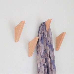 Puede incluir: Tres ganchos de pared de madera con forma de triángulo, con una bufanda estampada colgada en uno de los ganchos.