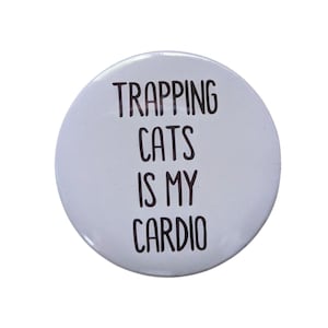 May include: A round, white button with black text that reads "TRAPPING CATS IS MY CARDIO". The text is centered and in a bold, sans-serif font. The button has a glossy finish.
