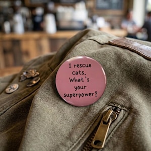 May include: A pink button with the text "I rescue cats. What's your superpower?" is pinned to a green jacket. The button is round and has a glossy finish. The jacket has a zipper and a brown leather detail.
