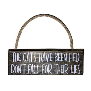 May include: A rustic wooden sign with a rope hanger reads "The cats have been fed. Don't fall for their lies."