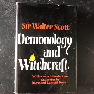 Op de afbeelding: Een zwart boek met de titel "Demonologie en Hekserij" van Sir Walter Scott. De titel is wit, de naam van de auteur oranje. Een gele vlam-afbeelding is zichtbaar. Bevat een inleiding en aantekeningen van Raymond Lamont Brown.