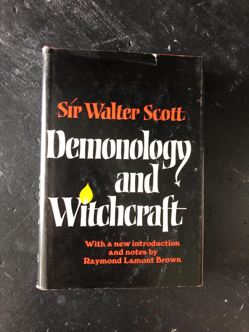 Op de afbeelding: Een zwart boek met de titel "Demonologie en Hekserij" van Sir Walter Scott. De titel is wit, de naam van de auteur oranje. Een gele vlam-afbeelding is zichtbaar. Bevat een inleiding en aantekeningen van Raymond Lamont Brown.
