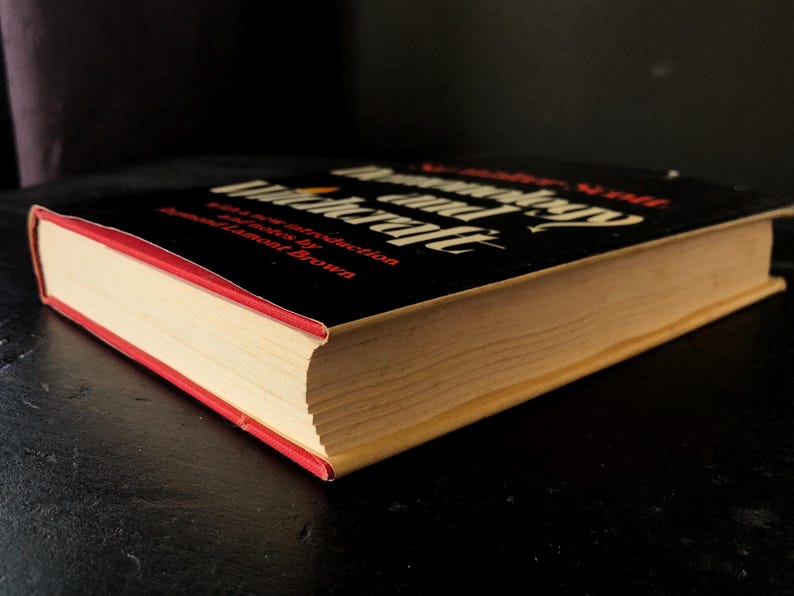 Op de afbeelding: Een hardcover boek getiteld "Demonology and Witchcraft" met een zwarte kaft en rode rand. De titel van het boek staat in witte en rode letters. De pagina's van het boek zijn vergeeld.