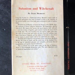 Op de afbeelding: De achterkant van een boek getiteld "Satanism and Witchcraft" van Jules Michelet. De omslag is gebroken wit met zwarte tekst en een zwarte rug. De tekst beschrijft de focus van het boek op middeleeuws bijgeloof en de Age of Darkness.