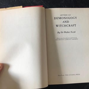 Op de afbeelding: Open boek getiteld "Letters on Demonology and Witchcraft" van Sir Walter Scott. De pagina's van het boek zijn gebroken wit met zwarte tekst. De kaft is rood en het boek is open op een blanco pagina.