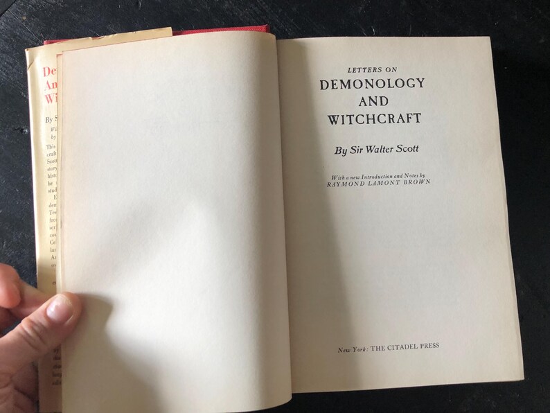 Op de afbeelding: Open boek getiteld "Letters on Demonology and Witchcraft" van Sir Walter Scott. De pagina's van het boek zijn gebroken wit met zwarte tekst. De kaft is rood en het boek is open op een blanco pagina.