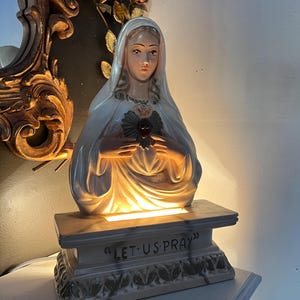 May include: A religious statue of the Virgin Mary, with a white veil and a heart on her chest. The statue is illuminated from within and sits on a base that reads "LET US PRAY".