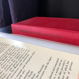 Op de afbeelding: Een rood hardcover boek getiteld "Demonology and Witchcraft" met gouden letters, rustend op open pagina's. De rug van het boek is zichtbaar en de pagina's zijn gebroken wit.