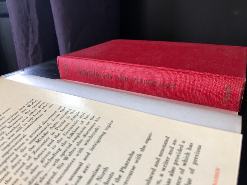 Op de afbeelding: Een rood hardcover boek getiteld "Demonology and Witchcraft" met gouden letters, rustend op open pagina's. De rug van het boek is zichtbaar en de pagina's zijn gebroken wit.