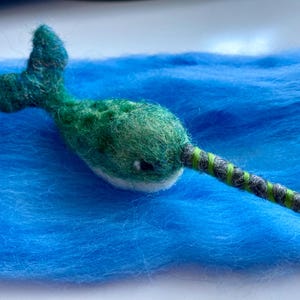 Puede incluir: Un narval de fieltro verde y blanco con un cuerno rayado negro y verde, descansando sobre una superficie de fieltro azul.