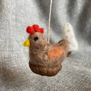 Könnte beinhalten: Handgefertigter Filz-Hühner-Ornament mit rotem Kamm und gelbem Schnabel. Das braune Huhn sitzt in einem gewebten Nest, mit einem orangefarbenen Akzent auf seinem Flügel. Im Hintergrund steht ein kleiner weißer Baum mit rosa Perlen.