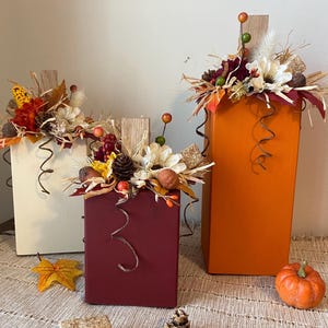 May include: Three rectangular wooden decorative boxes in cream, burgundy, and orange, adorned with fall-themed floral arrangements. Each box has a wooden accent and decorative wire. A small pumpkin and fall leaves are also present.