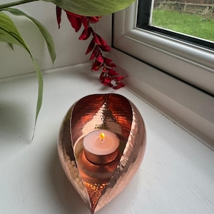 May include: A teardrop-shaped candle holder crafted from hammered copper with a metallic finish. The holder features a curved, open design that cradles a single lit white tea light candle in its centre, reflecting the warm glow on its surface.