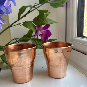 May include: Two matching drinking cups crafted from polished copper, each featuring a distinct hammered texture across the exterior surface. The cups have a slightly tapered cylindrical shape with a smooth, rounded rim at the top edge.