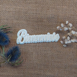 Op de afbeelding: Een wit, gelaagd woordkunstwerk in cursieve stijl met de tekst "Bienenwiese". Het stuk staat tegen een getextureerde, bruine jute achtergrond, met blauwe distel en witte bloemen in de hoeken.