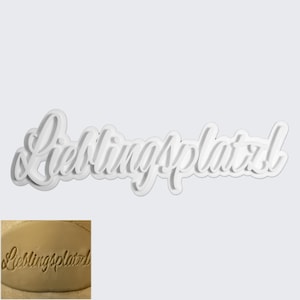Op de afbeelding: Een witte, gelaagde koekjessnijder in een cursief lettertype dat "Lieblingsplatz" spelt. De afbeelding toont ook een kleine, ingezette afbeelding van hetzelfde woord dat in een lichtbruin oppervlak is geperst, waarschijnlijk deeg of klei.