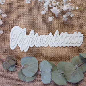 Op de afbeelding: Witte plastic koekjesvormer met de woorden "Töpferwerkstatt" in een cursief schrift.